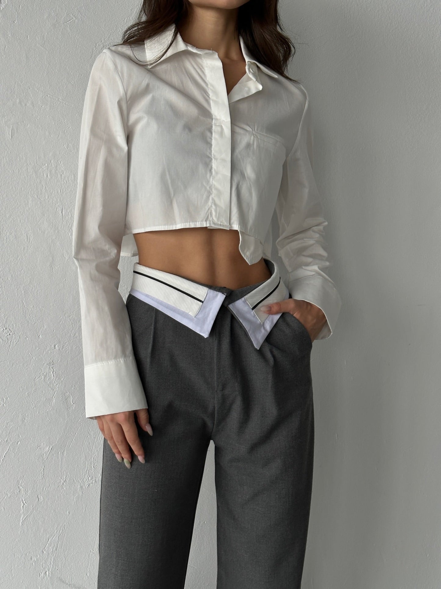 A person models a stylish outfit featuring a white cropped blouse paired with the elegant Rina Pants, high-waisted gray trousers with a unique double waistband and wide-leg design, crafted from breathable fabric, against a plain background.