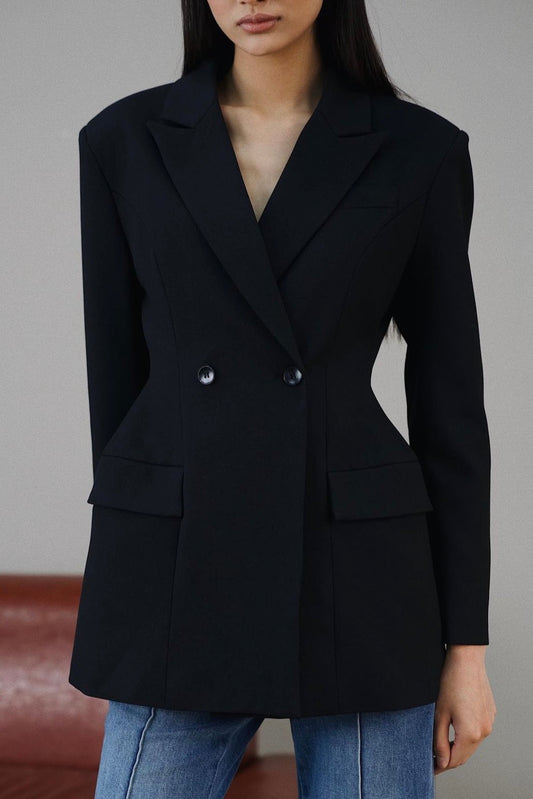 Dressed in the Olivia tailored blazer with structured shoulders and wide lapels, paired with blue jeans, a person stands in the room with their face obscured.
