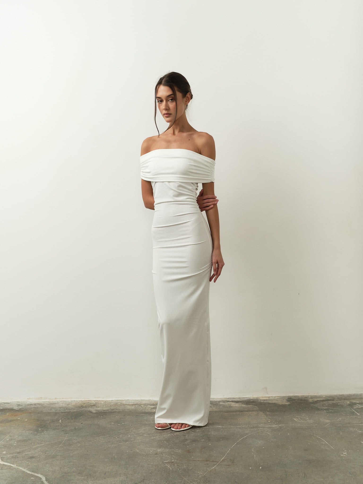A woman stands against a plain white background, wearing the Scarlett Dress, an off-the-shoulder, form-fitting gown that reaches the floor. The dress perfectly highlights her feminine silhouette. Her hair is styled up, and she stands with her hands gently clasped together.